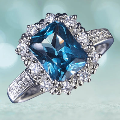 Marselles Ring featuring a large rectangular blue gemstone surrounded by smaller clear stones, set in rhodium-plated sterling silver, designed for women’s jewelry collection.