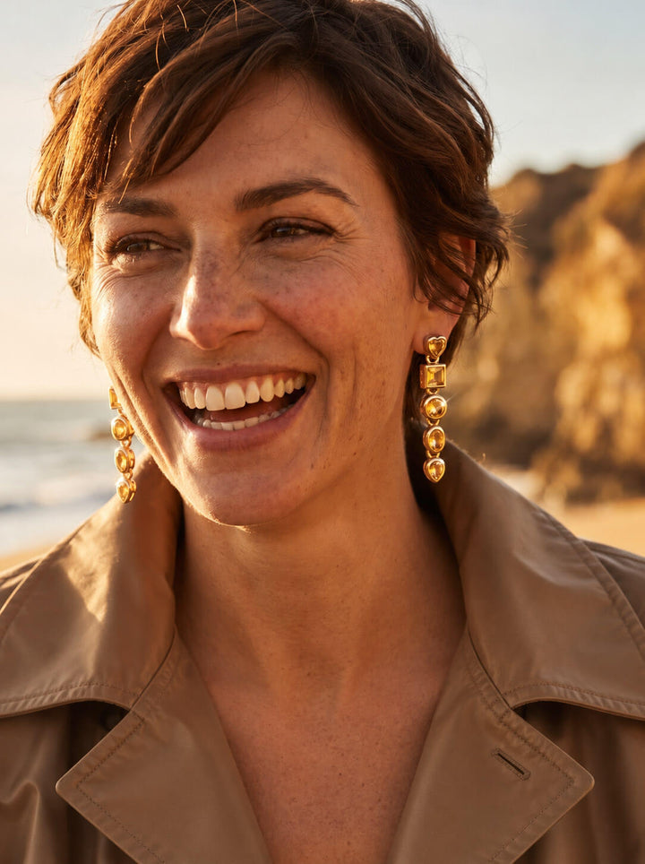 Citrine Symphony Earrings with golden drop settings and sparkling round citrine stones worn by a smiling woman on a sunlit beach