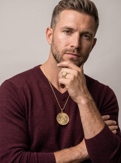 Man wearing a gold American Glory Pendant necklace and matching gold ring, dressed in a burgundy sweater, posing thoughtfully against a neutral background.