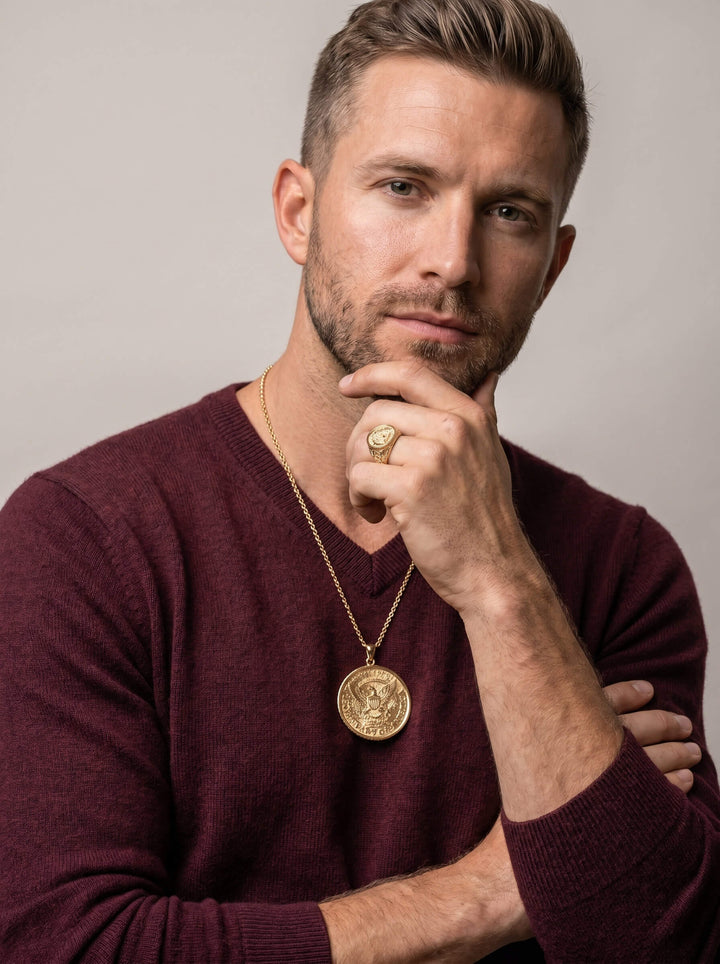 Man wearing a gold American Glory Pendant necklace and matching gold ring, dressed in a burgundy sweater, posing thoughtfully against a neutral background.