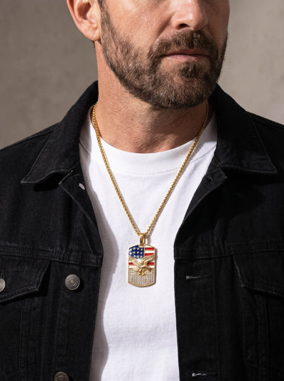 Man wearing a gold chain necklace featuring the Patriot’s Crest Pendant with an American flag design and an eagle emblem, styled with a white shirt and black jacket.