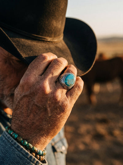 Close-up of a weathered hand wearing a Turquoise Frontier Ring with a large blue stone, alongside a beaded turquoise bracelet, set against a rustic outdoor background.