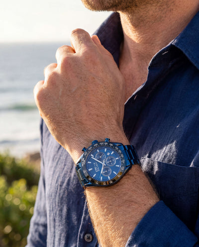 Close-up of a man wearing a Midnight Velocity Men's Watch with a blue metal band and dial, showcasing a sporty and elegant timepiece by the ocean.