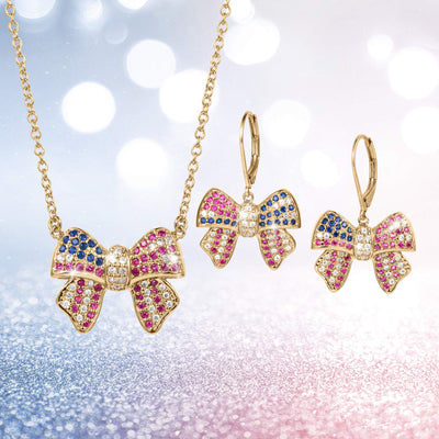 Gold-tone bow-shaped necklace and matching earrings adorned with pink, blue, and white stones from the Heritage Bow Collection, displayed on a sparkling background