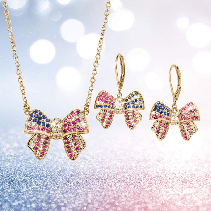 Gold-tone bow-shaped necklace and matching earrings adorned with pink, blue, and white stones from the Heritage Bow Collection, displayed on a sparkling background
