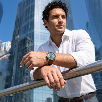 Man wearing a Legacy Chrono Men's Watch with a brown leather strap, posing confidently against a backdrop of modern glass skyscrapers, showcasing stylish timepieces.