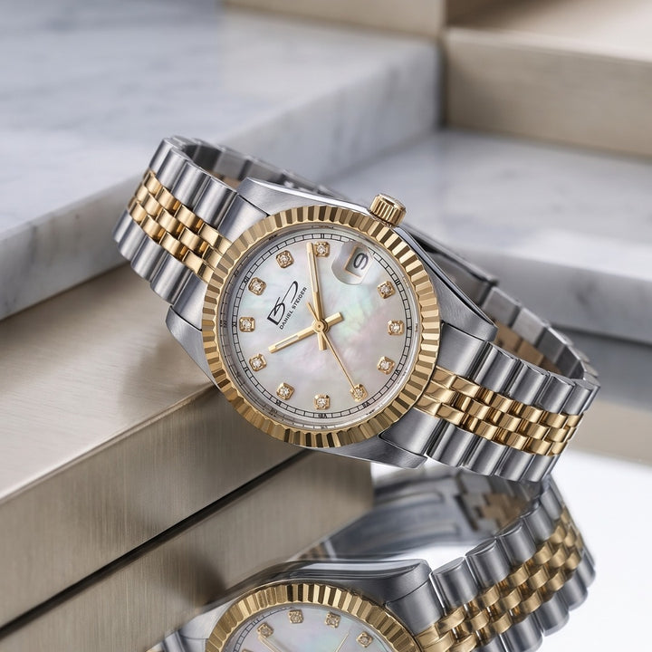 A two-tone stainless steel and gold wristwatch with a white mother-of-pearl dial, crystal hour markers, and a date window, displayed on a metallic surface with a marble background. Daniel Steiger ladies Kudos