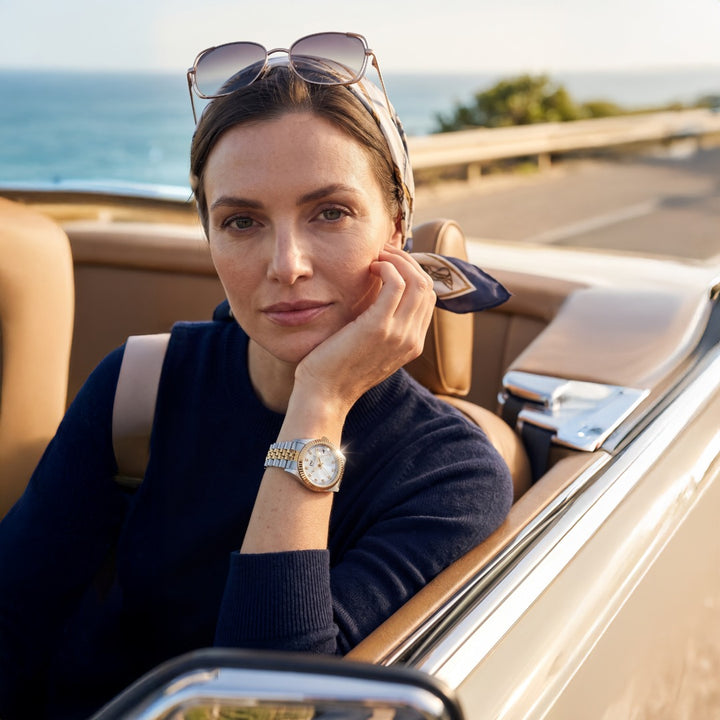 A woman sitting in a beige convertible car by the ocean, wearing a silver-toned watch with a white dial and a navy headscarf with sunglasses resting on her head. Daniel Steiger ladies Kudos