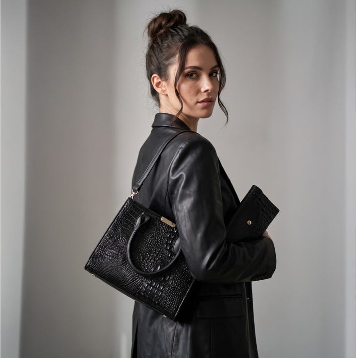 A woman in a black leather jacket carries a black crocodile-embossed leather handbag and matching clutch against a neutral gray background. Gabriella Bag Collection featuring a black crocodile texture handbag wallet and mini purse with polished gold accents Up To 75% Off Retail