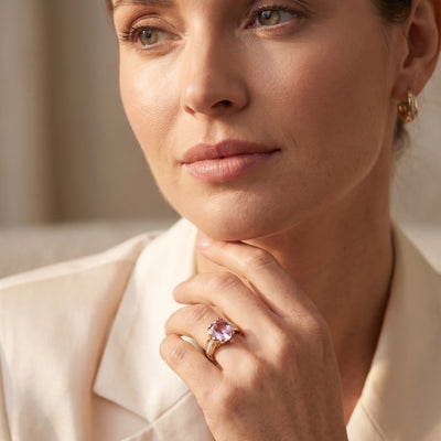 Close up portrait of a woman resting her hand beneath her chin while wearing a gold ring with a purple gemstone and matching gold earrings